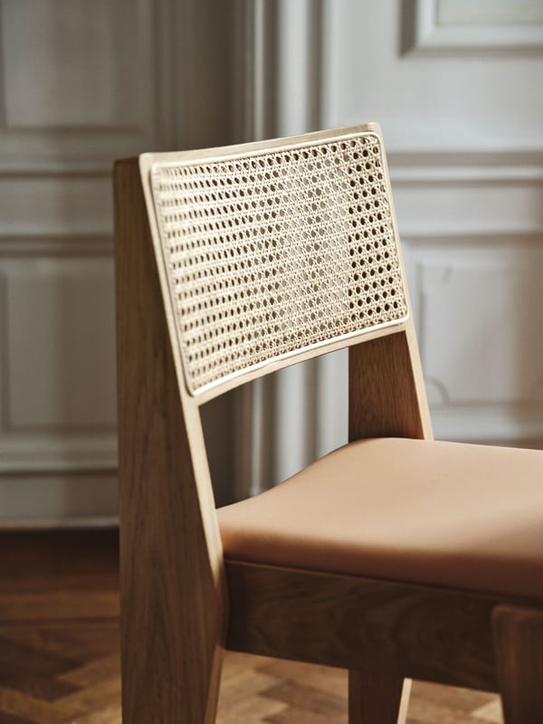 A STOCKHOLM 2025 chair with a solid oak frame, leather seat, and rattan backrest stands on parquet flooring.