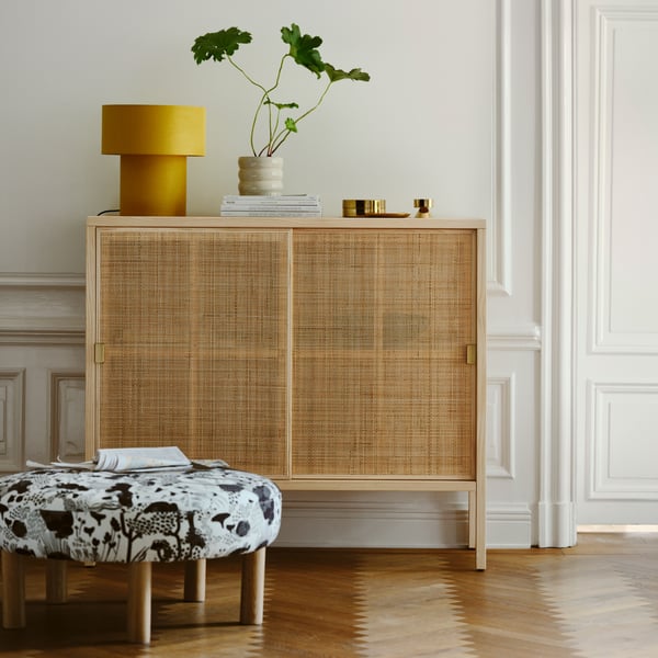 A STOCKHOLM 2025 cabinet with sliding doors in rattan and pine on parquet, with a table lamp on top and a pouffe in front.